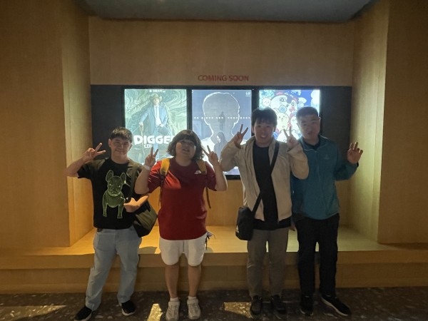 동아리원 4명이 나란히 서서 영화포스터 3개가 붙여진 곳 앞에서 브이를 하고 포즈를 취하고 있습니다.
