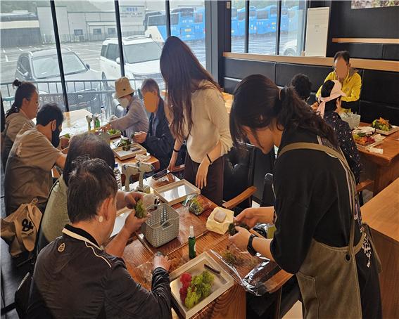 9명의 사람들이 스칸디아모스 풀을 만지고 있다. 강사는 일어나서 설명하고 있고 다른 사람들은 4명씩 사각테이블에 앉아있다