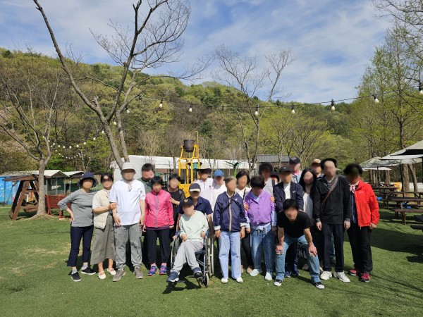30대에서 50대 사이의 장애인 참여자 11명과 활동지원사 11명이 함께 찍은 단체사진으로, 화창한 날씨 속에서 파란 하늘과 녹색의 인조잔디, 연두빛 잎사귀가 돋보이는 나무들이 배경을 이루며 봄의 산뜻한 분위기를 전합니다.