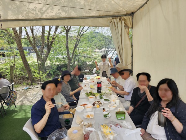 10명이 앉을 수 있는 큰 테이블에 상추, 고추, 양파, 비빔면, 그리고 맛있게 익은 고기를 놓고 참여자들과 활동지원사들이 즐겁게 먹고 있으며, 몇 명은 카메라를 바라보며 브이 포즈를 취하고 있다.