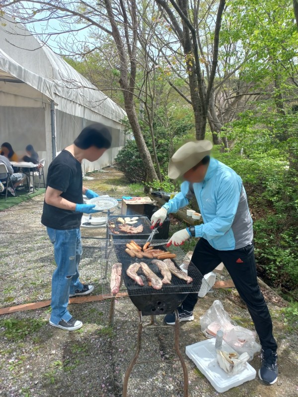 두 명의 성인 남성이 숯불 그릴에 삼겹살과 소세지, 버섯을 굽고 있는 모습이다.
