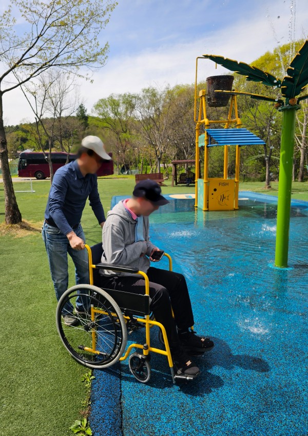 남성 활동지원사가 휠체어를 타고 있는 남성 참여자에게 발목까지 오는 깊이의 물 놀이터에서 분수가 나오는 곳에서 물을 느끼도록 돕고 있는 모습이다.